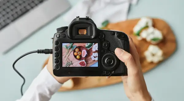 Dłoń trzyma aparat fotograficzny, na którego ekranie widać zdjęcie jedzenia.